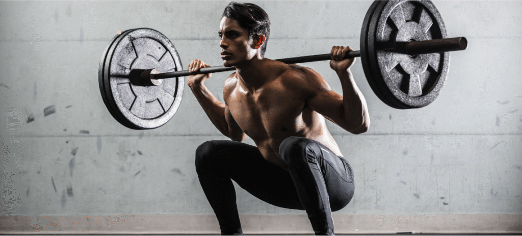 Muscular man performing barbell back squat with heavy weights in gym, demonstrating proper strength training form and technique